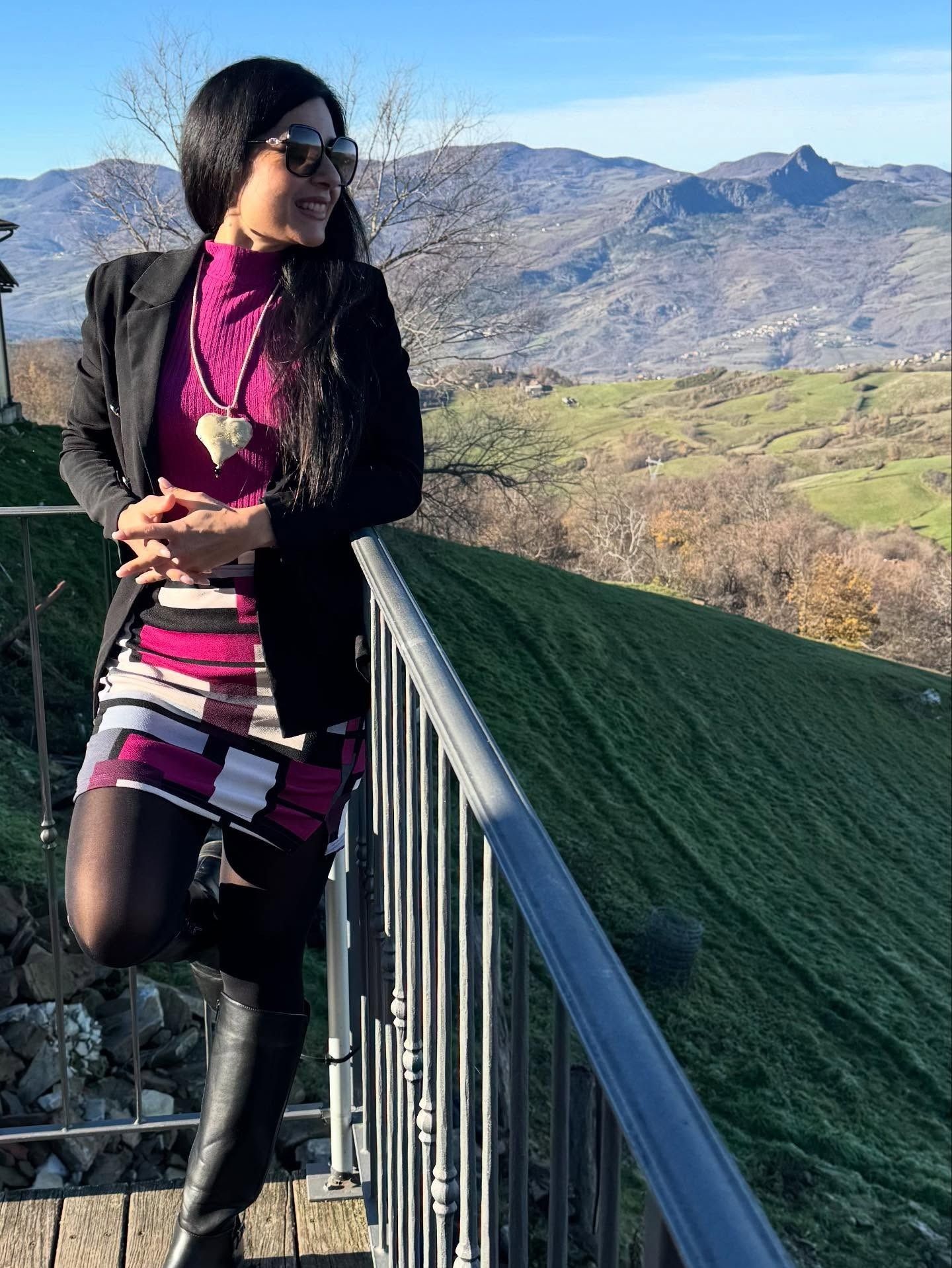 Una persona che indossa un blazer, un vestito e degli stivali si appoggia alla ringhiera di un balcone con vista su un paesaggio montuoso.