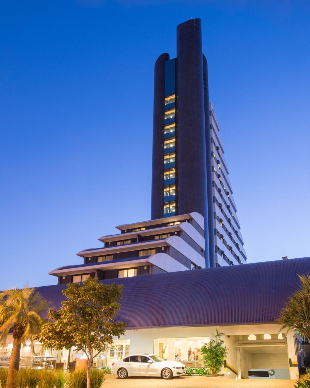 Exterior do hotel ao anoitecer, com uma grande placa, entrada bem iluminada e área gramada.