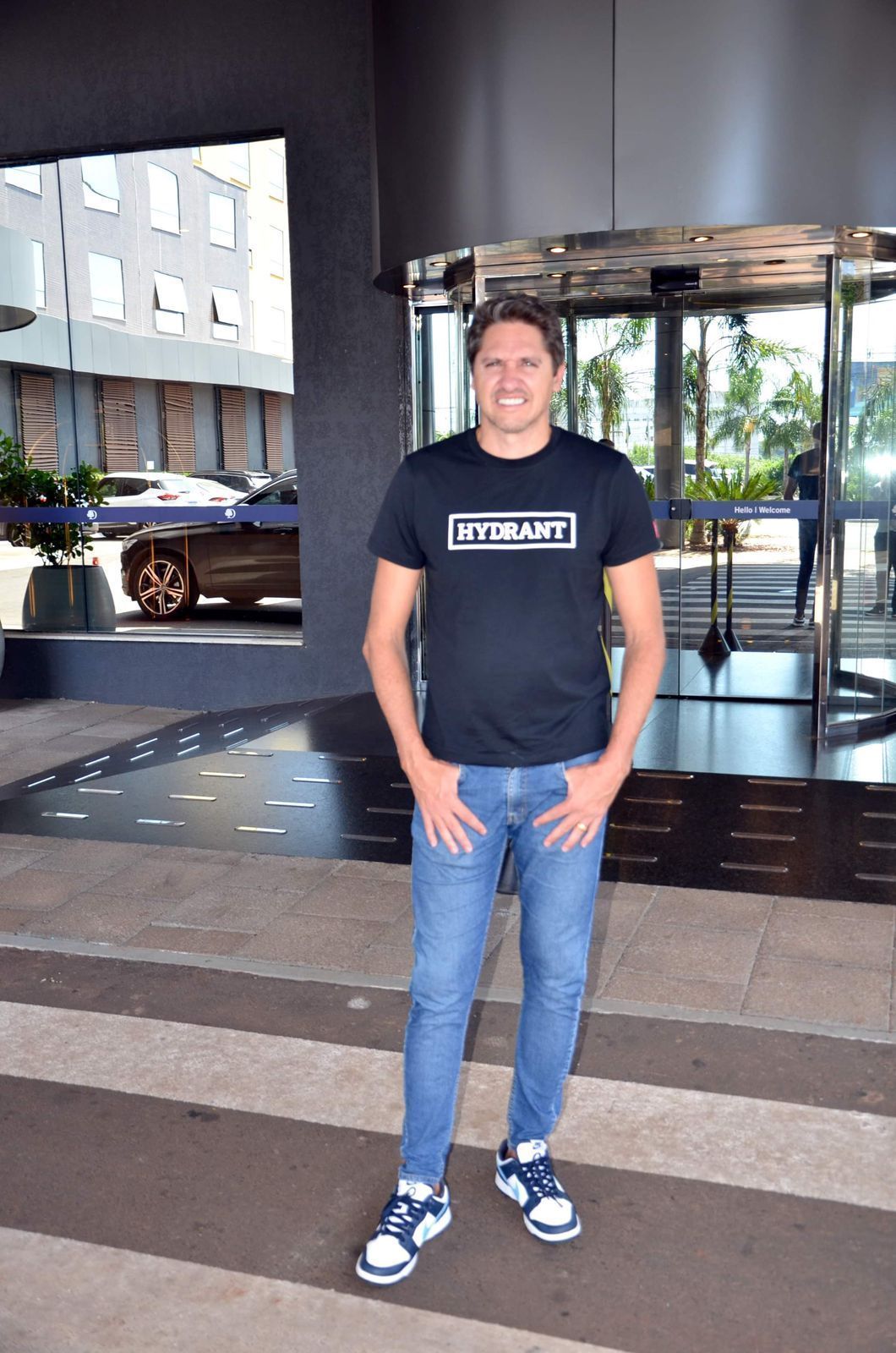 Homem de camiseta preta e jeans está do lado de fora de um prédio com as mãos nos bolsos e sorrindo.