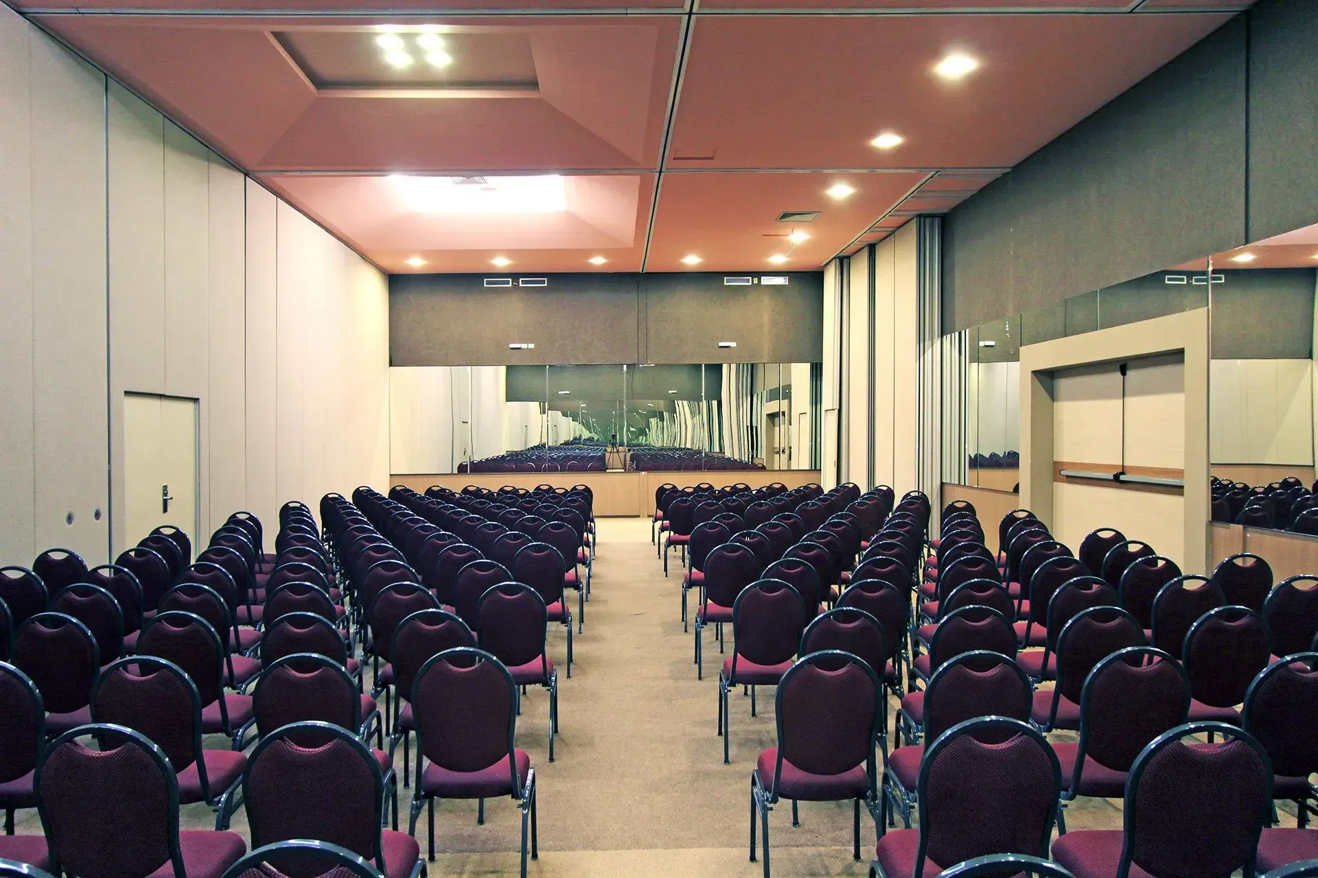 Auditório com fileiras de cadeiras cor de vinho de frente para um palco, carpete e paredes bege, luzes no teto.