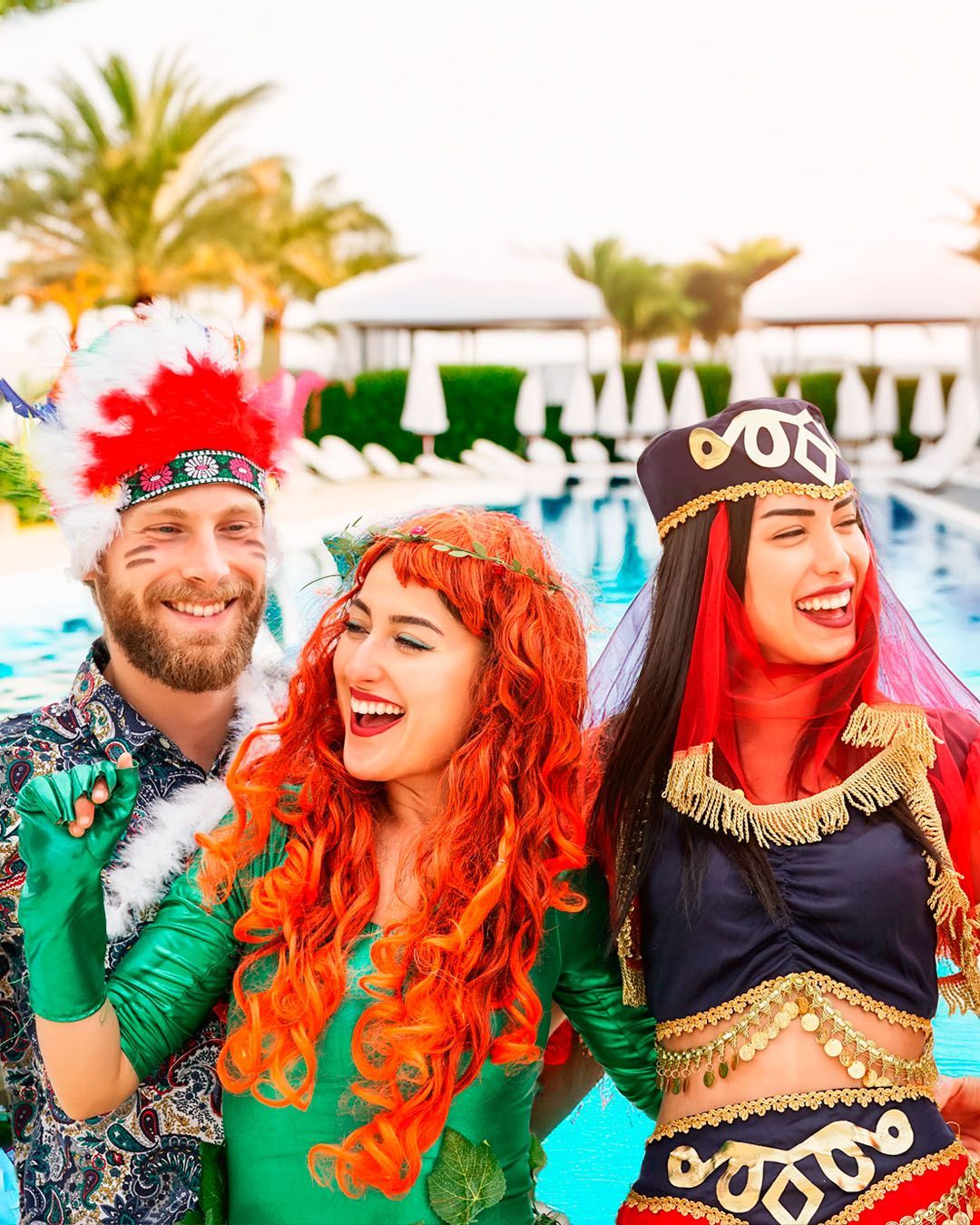 Três pessoas fantasiadas perto de uma piscina, rindo. Uma usa um cocar indígena, outra uma roupa de hera venenosa e a terceira uma roupa de dança do ventre. Carnaval 2026.