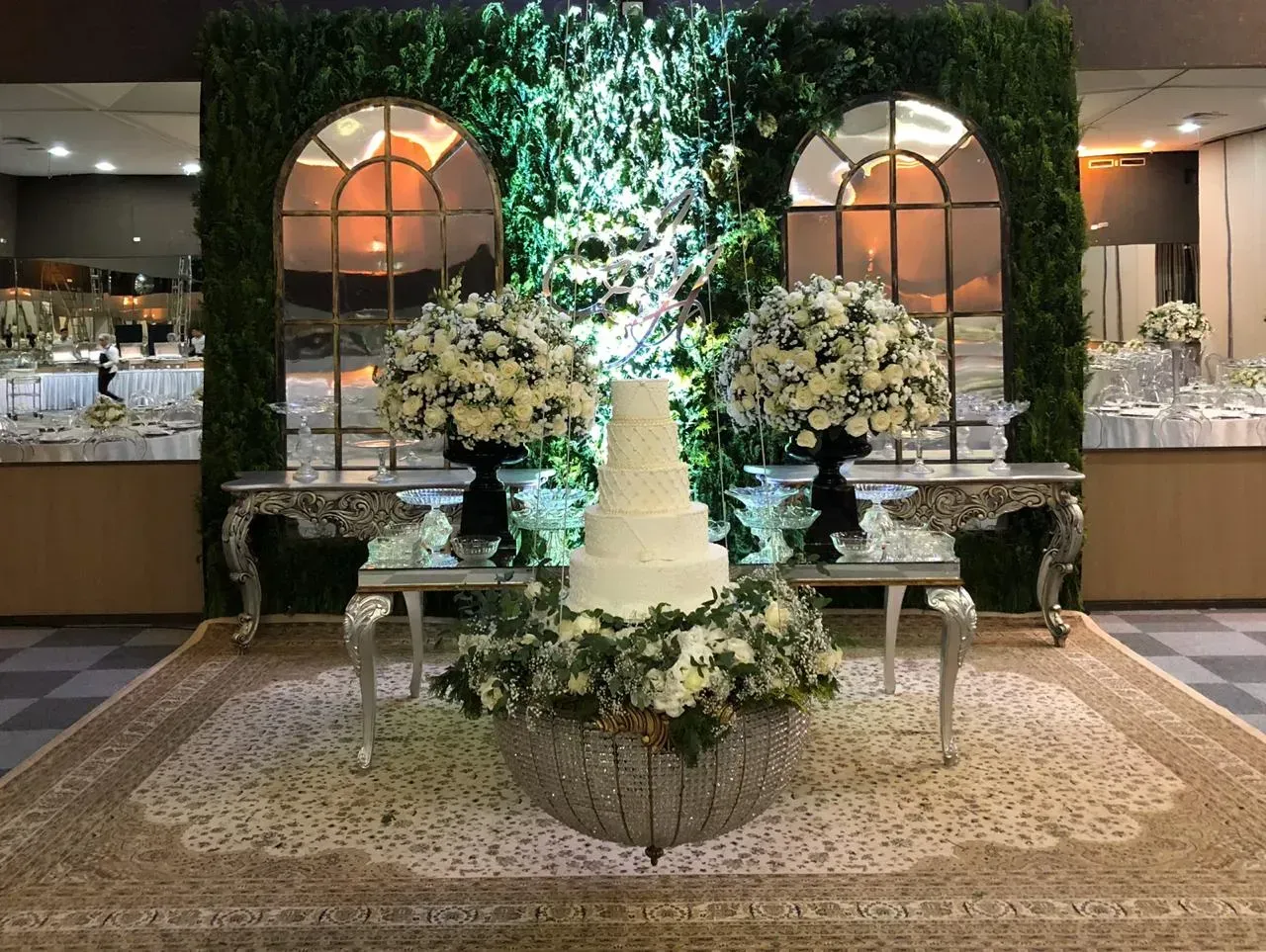 Exibição de bolo de casamento com flores, espelhos e folhagens em um ambiente formal.