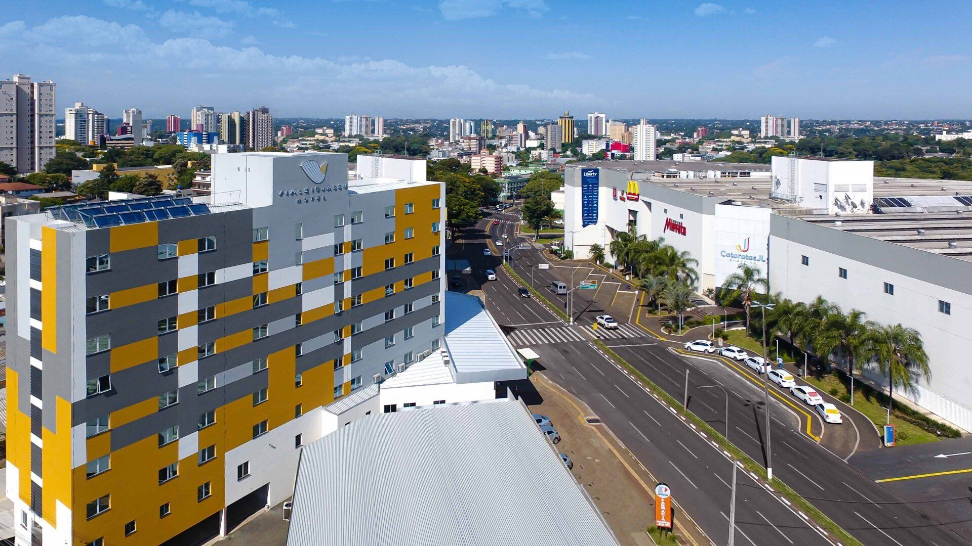 Paisagem urbana com um prédio de vários andares com painéis amarelos e cinza; estradas, lojas e outros edifícios são visíveis.