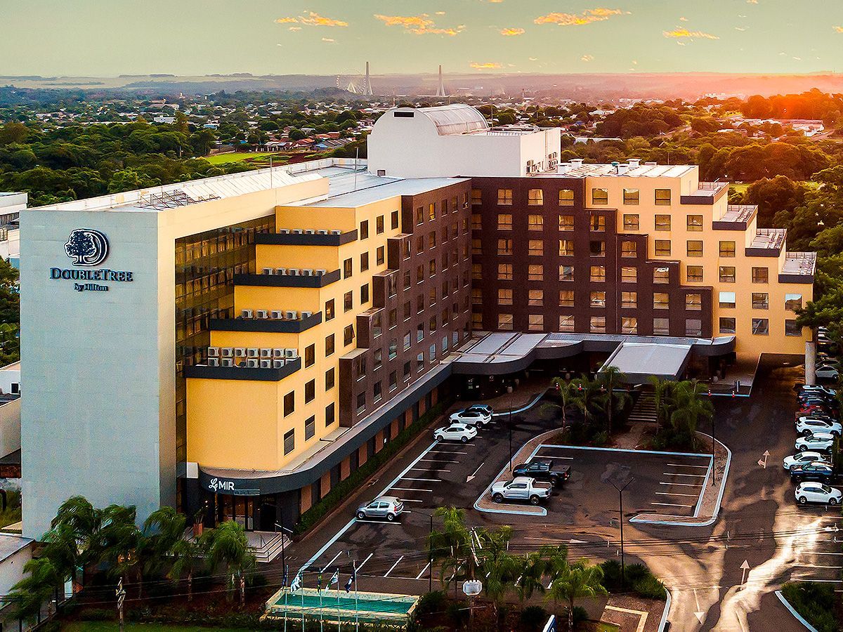 Vista aérea do hotel DoubleTree by Hilton, em tons de amarelo e marrom, ao pôr do sol, com o estacionamento e as árvores ao redor.