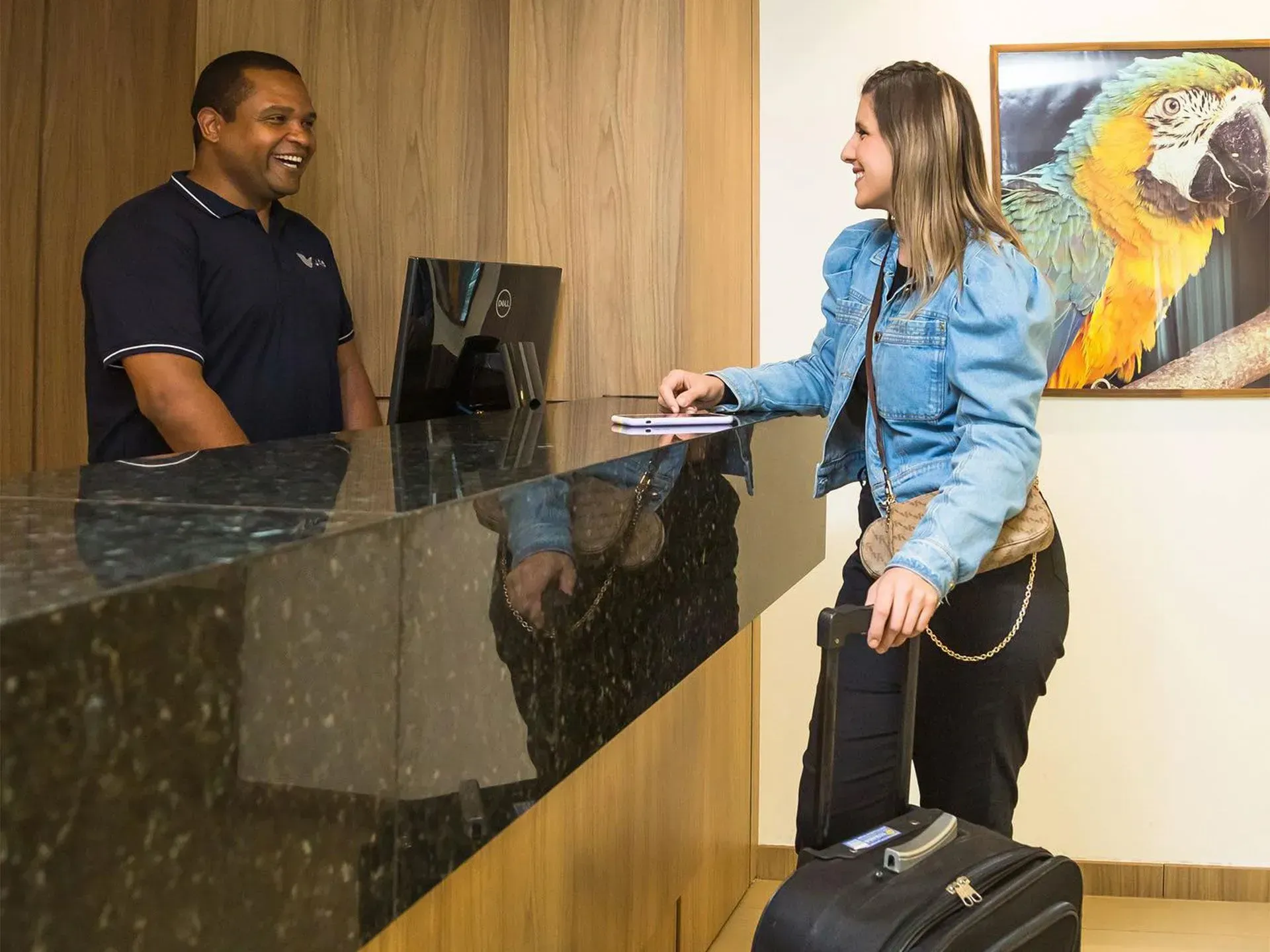 Recepcionista de hotel auxiliando uma mulher com a bagagem. Ambos sorrindo para a recepção. Pintura de um papagaio na parede.