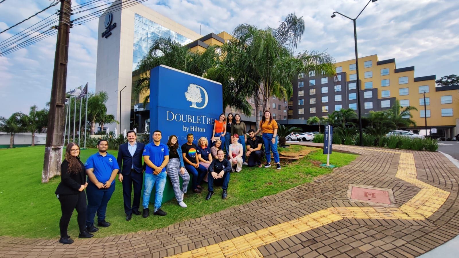 Grupo de pessoas em frente à placa de um hotel DoubleTree. Grama verde, céu azul e uma estrada de tijolos são visíveis.