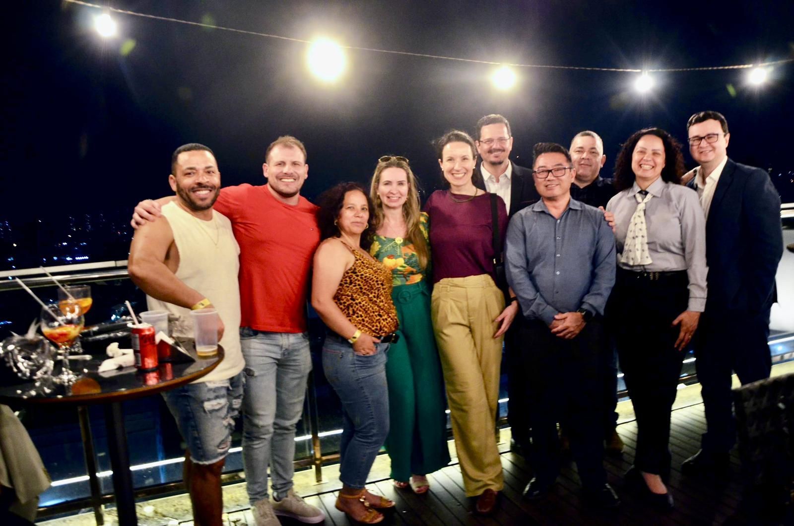 Grupo de pessoas sorrindo para uma foto no terraço de um prédio à noite, coquetéis na mesa e luzes da cidade ao fundo.