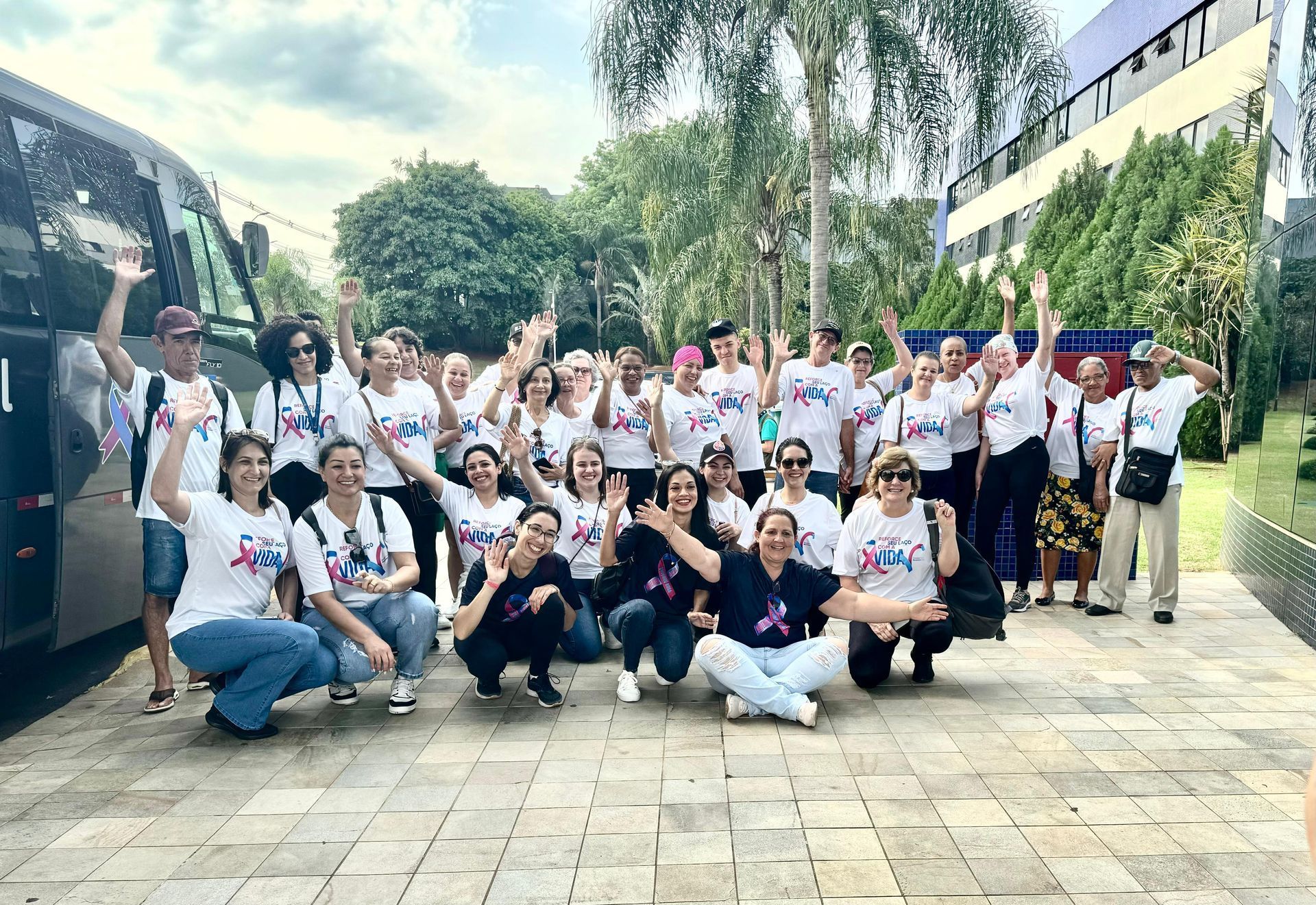 Grupo de pessoas acenando, vestindo camisas brancas, perto de um ônibus e um prédio.