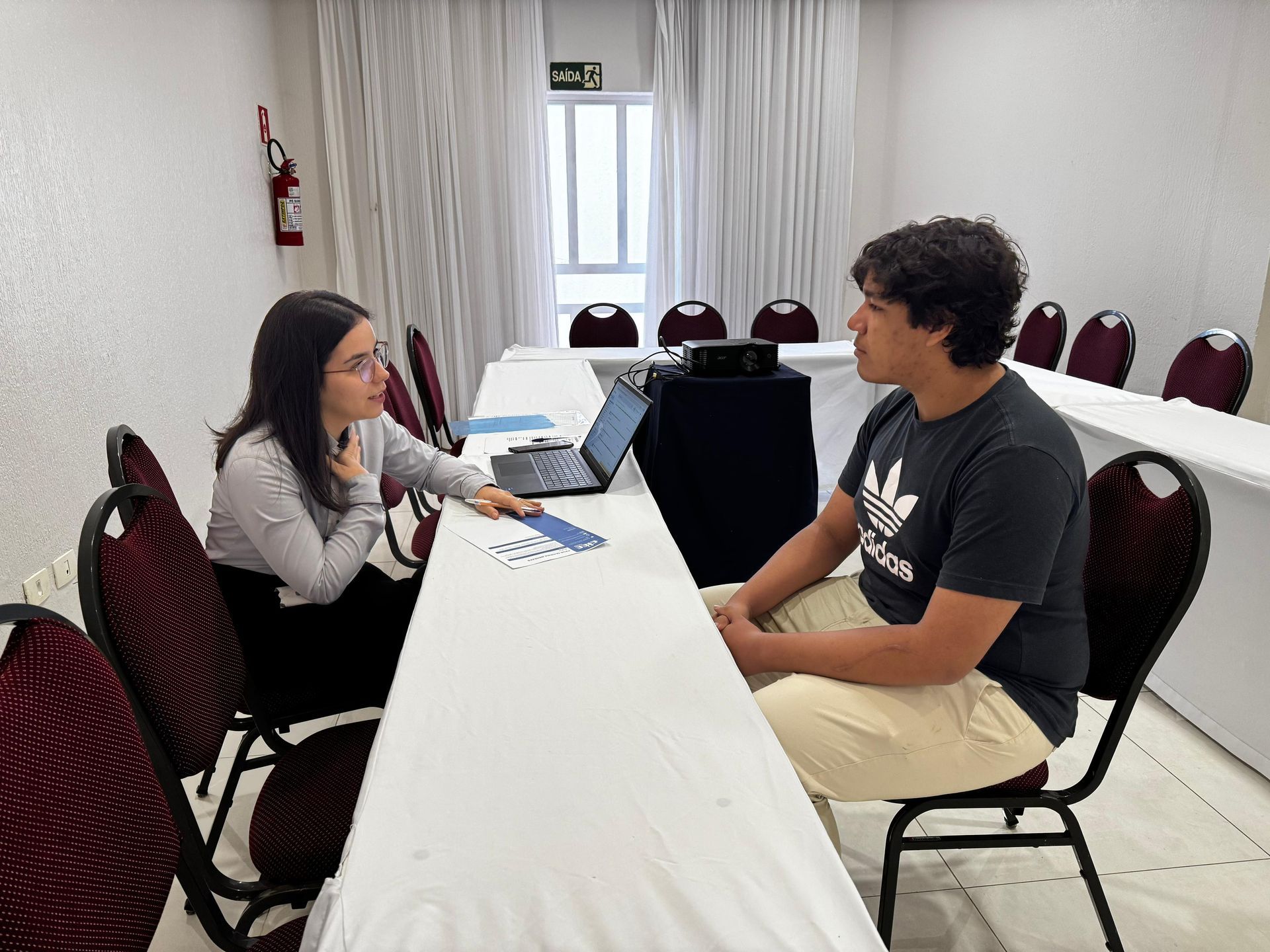 Mulher entrevistando um homem em uma mesa, em uma sala com uma janela e várias cadeiras.