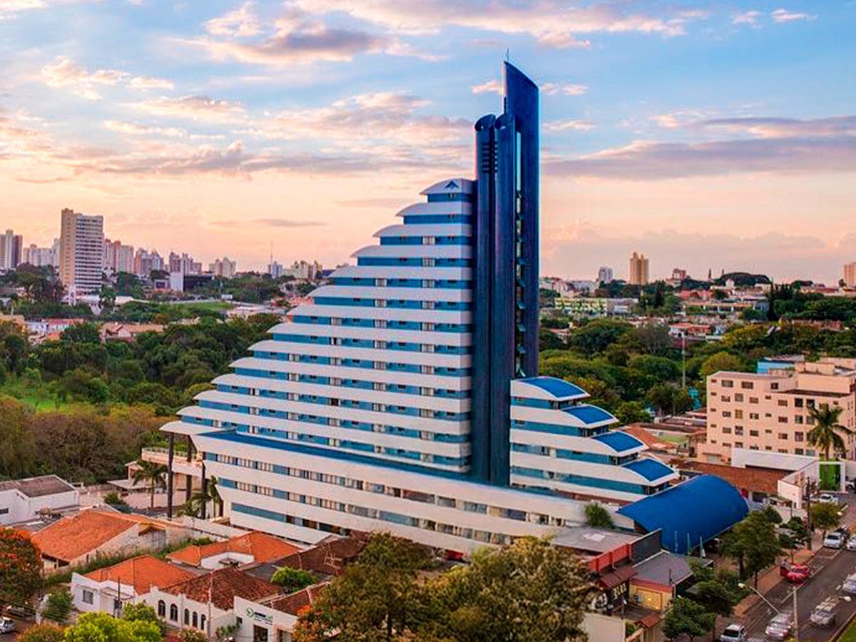 Um prédio de hotel alto, moderno e escalonado, com detalhes em azul e branco, destaca-se contra um céu ao nascer do sol em uma paisagem urbana.