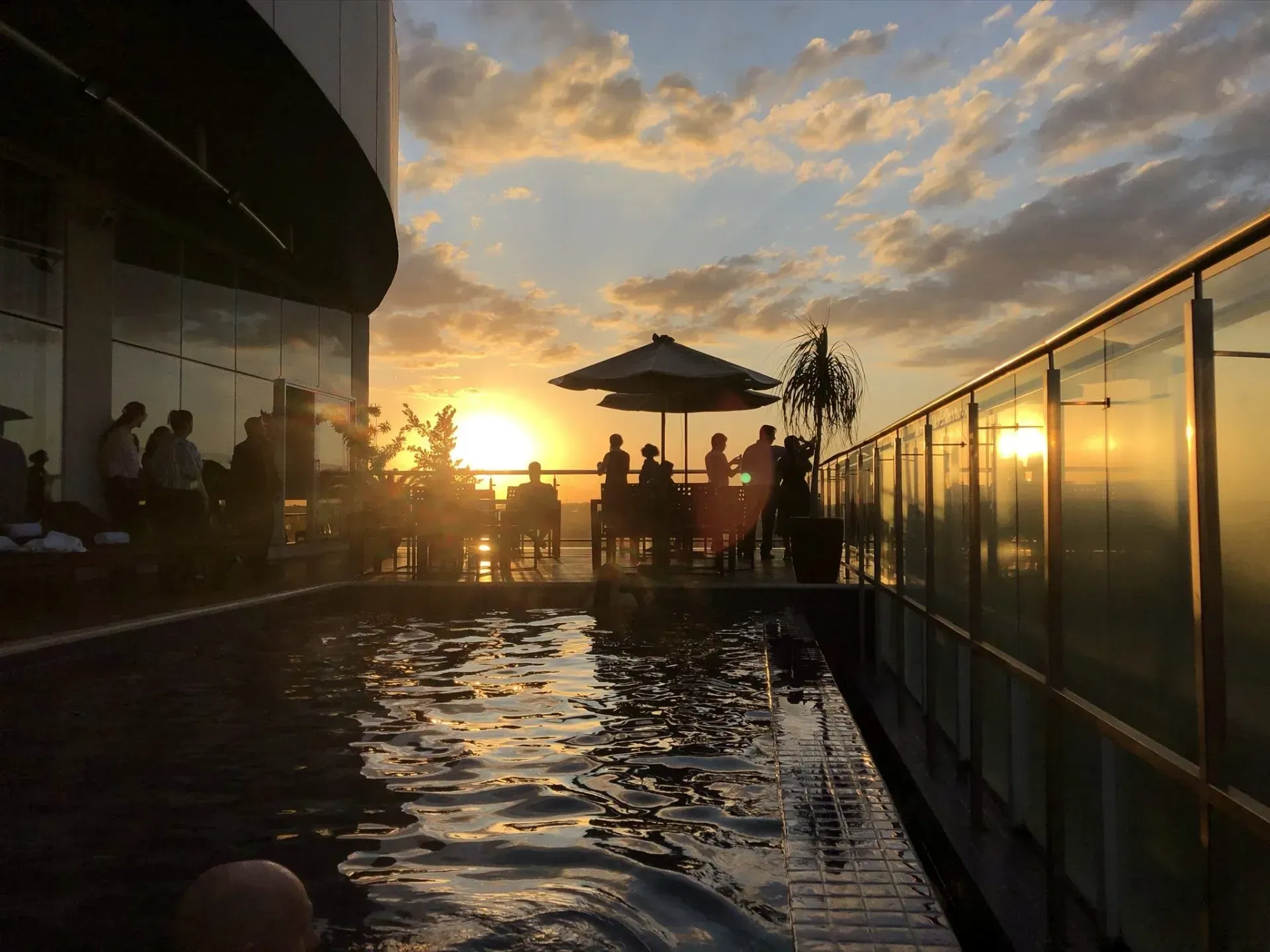 Piscina na cobertura ao pôr do sol com silhuetas de pessoas perto de uma mesa. A água reflete a luz dourada do sol.