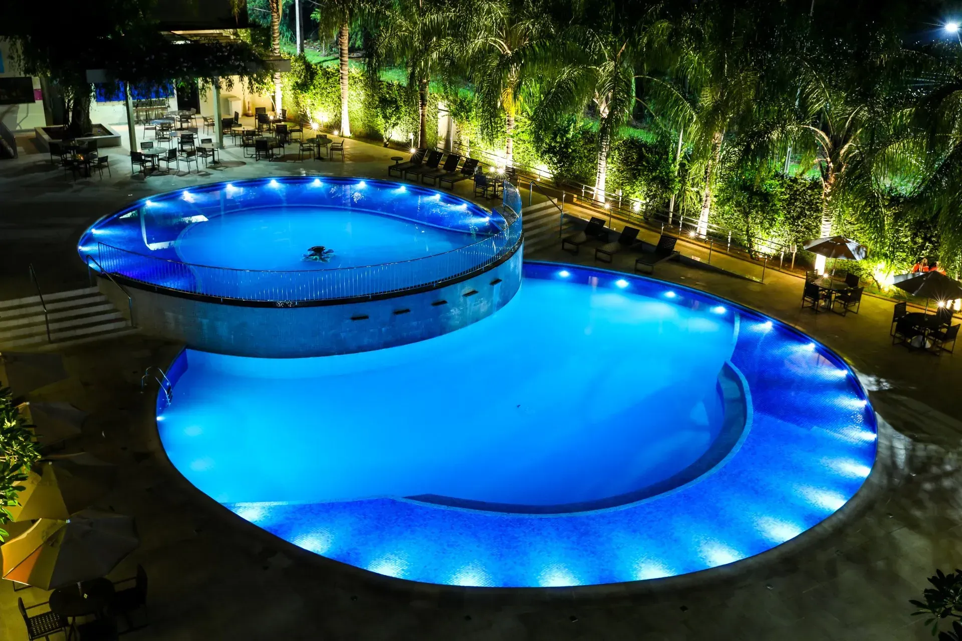 Vista noturna de uma piscina iluminada em azul com uma banheira de hidromassagem circular acima dela, cercada por árvores.