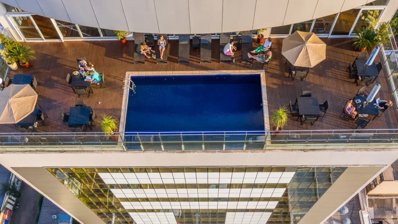 Piscina na cobertura com espreguiçadeiras e guarda-sóis; pessoas relaxando ao sol. Vista da cidade abaixo.