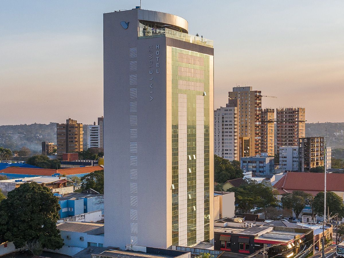 Um edifício alto, moderno e de cor clara, com fachada de vidro espelhado, destaca-se na paisagem urbana ao pôr do sol.