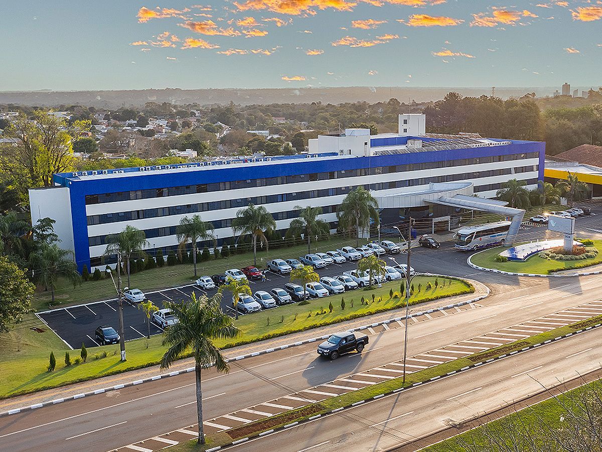 Um hotel comprido, de vários andares, com fachada branca e azul, localizado ao lado de uma rua com um estacionamento cheio de carros.