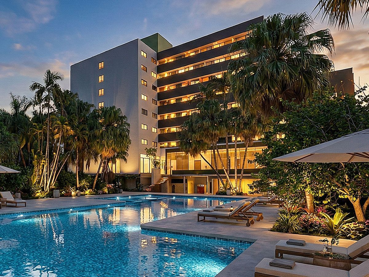 Um moderno hotel resort com janelas iluminadas oferece vista para uma piscina turquesa rodeada por palmeiras tropicais.