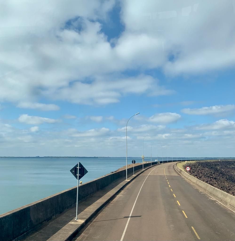 Estrada sobre uma represa com água azul à esquerda, céu azul com nuvens.