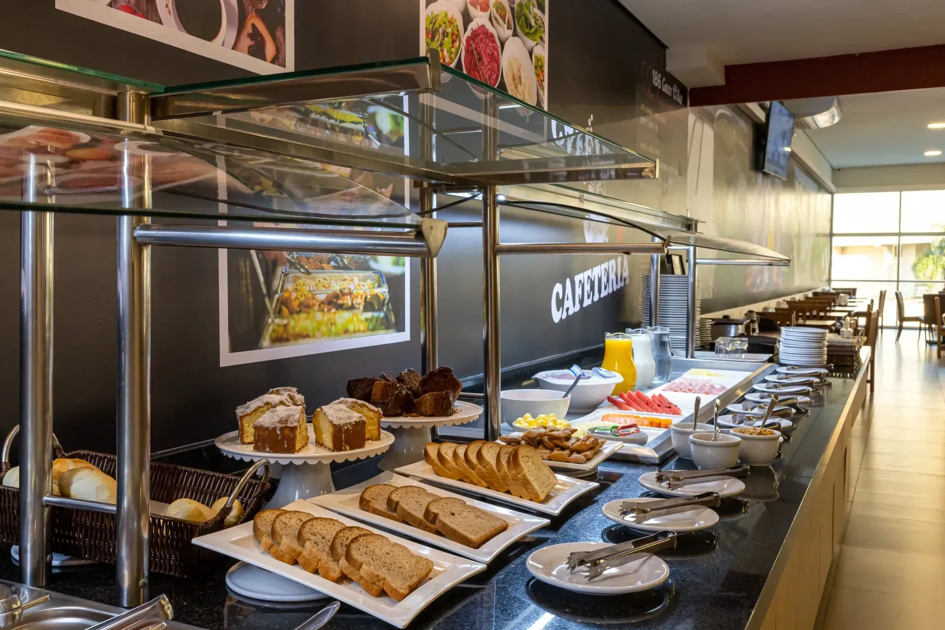 Buffet de café da manhã com diversos itens alimentares: pães, bolos, frutas e pratos quentes, em um refeitório.