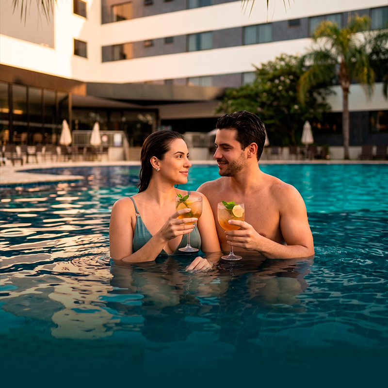 Duas pessoas brindam com coquetéis e sorriem perto de um prédio do resort ao pôr do sol, na piscina de um hotel.