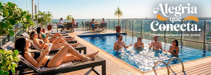 People enjoying rooftop pool, some swimming, some sunbathing, others sitting at tables. 