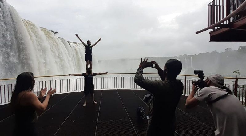 Acrobatas fazendo uma pirâmide humana perto de uma cachoeira; pessoas os fotografam em um deck.