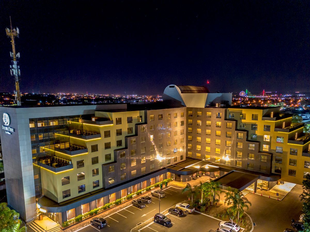 Visão noturna de um hotel com janelas iluminadas, um estacionamento e luzes da cidade ao fundo.