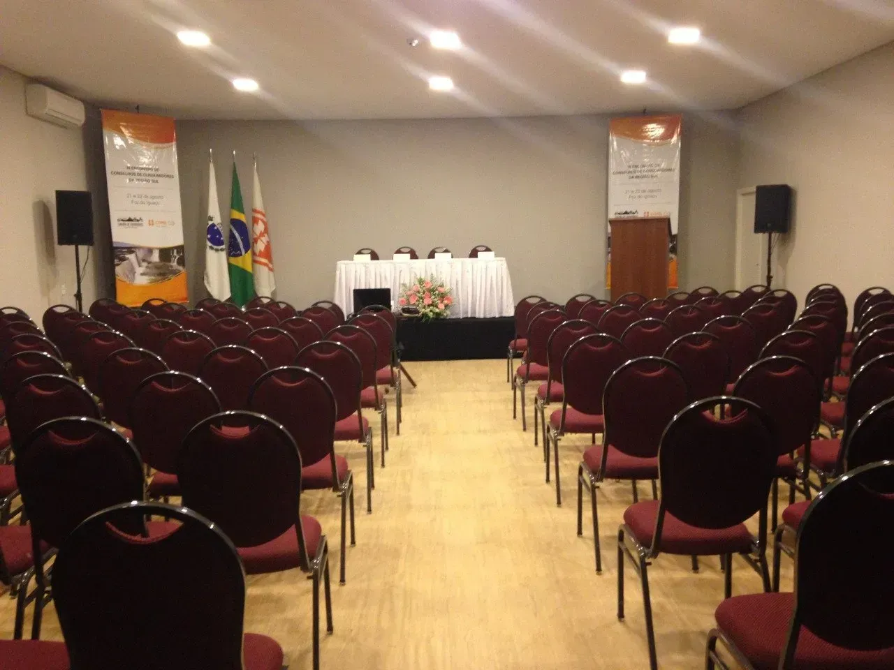 Sala de conferências com fileiras de cadeiras vermelhas de frente para um palco com bandeiras e um pódio.