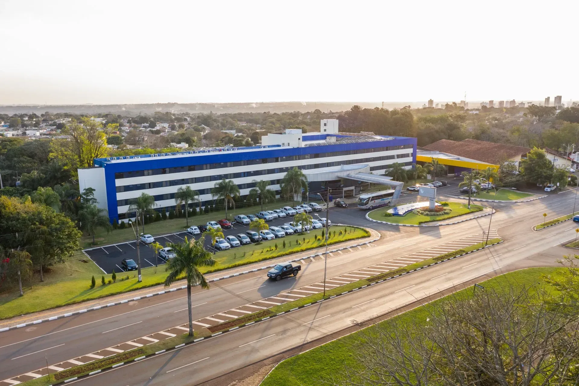Grande edifício azul e branco, provavelmente um hospital ou escritório, cercado por estradas, árvores e carros estacionados.