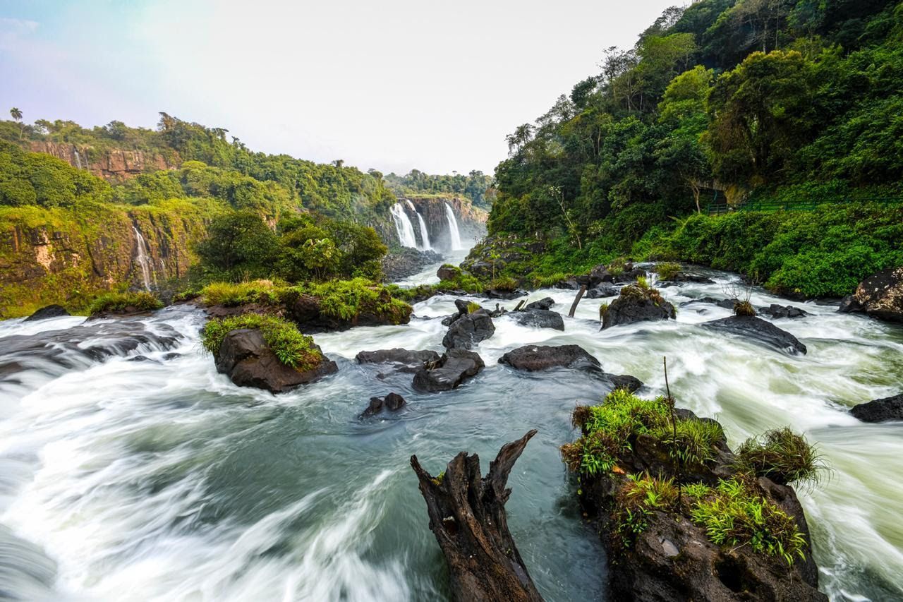 Cachoeira desaguando em um rio rochoso, cercada por árvores verdejantes.