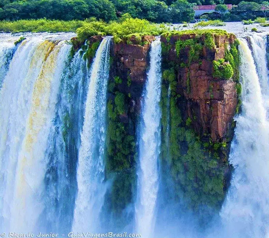 Cachoeira caindo sobre um penhasco rochoso, com folhagem verde no topo. Água azul e branca.