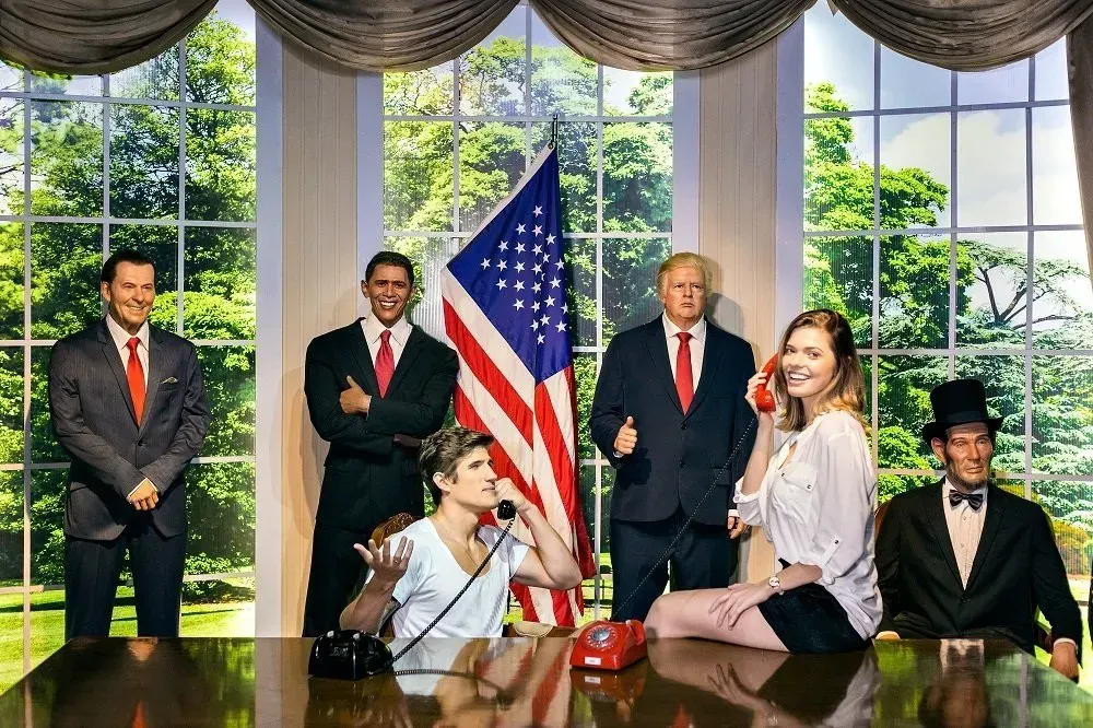 Figuras de cera: Presidentes, funcionários e uma mulher ao telefone no Salão Oval. Uma bandeira americana no centro.