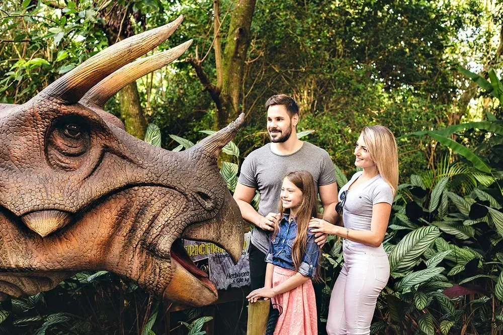 Família posa perto de escultura de dinossauro em ambiente verdejante.