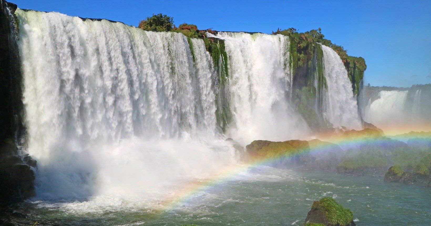 Cachoeira caindo sobre rochas, com um arco-íris nos respingos, sob um céu azul.