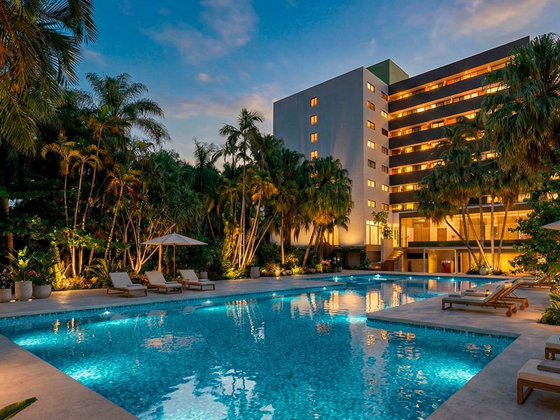 Uma grande piscina de hotel iluminada ao entardecer, rodeada por palmeiras exuberantes e espreguiçadeiras, com o edifício ao fundo.