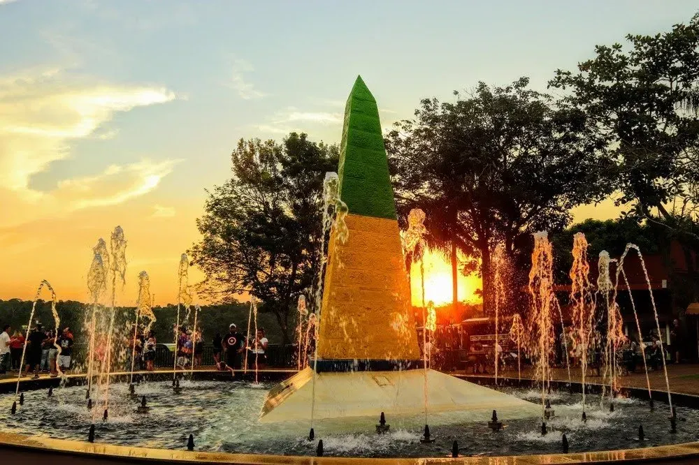 Fonte com jatos d'água jorrando para cima ao redor de uma escultura central. O pôr do sol lança um brilho alaranjado.