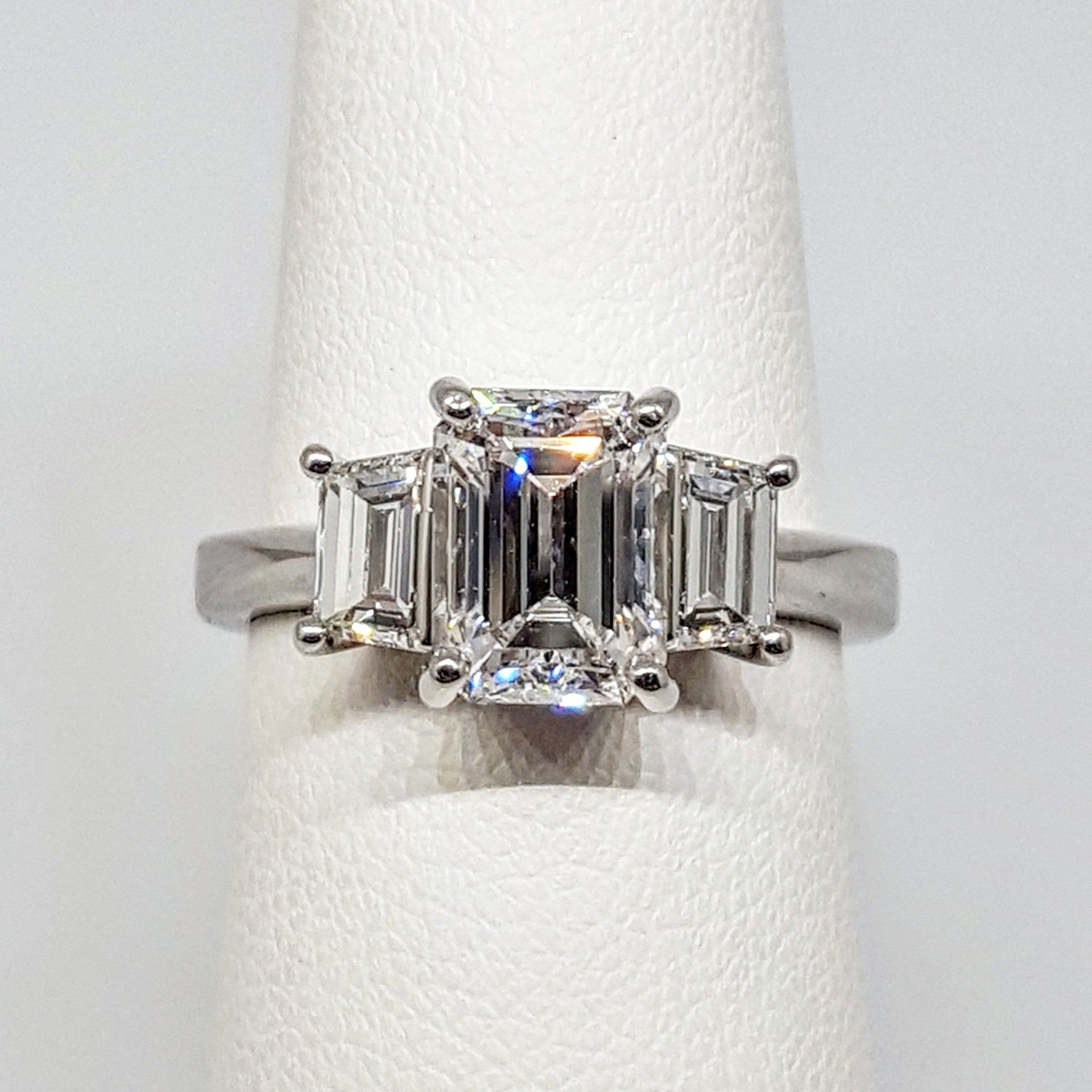 A white gold ring with three emerald cut diamonds on a finger.