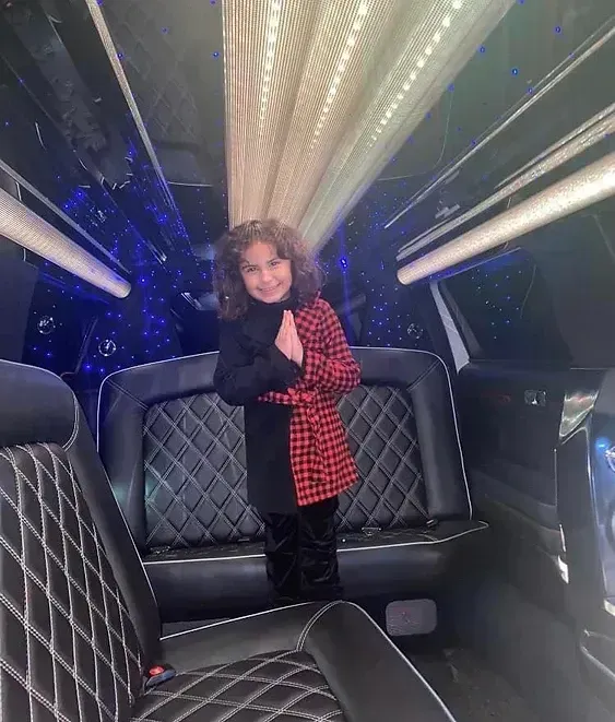 Young child in a limo, standing, wearing a red and black jacket. Interior with decorative lights and black seats.