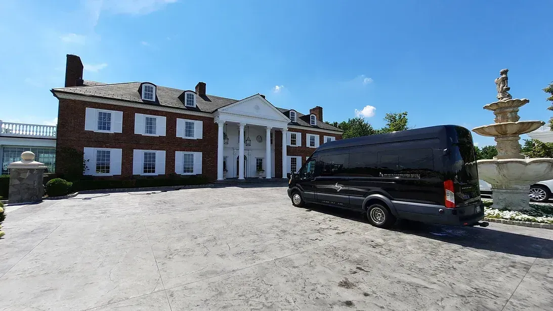 Black van parked in front of a brick mansion with white columns and a fountain on a sunny day.