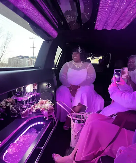 Woman in white dress smiles in a limo with purple lighting. Others take photos.