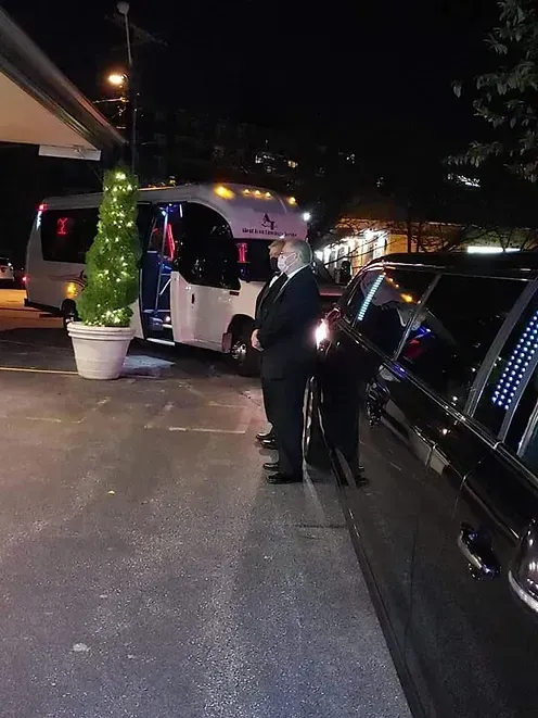 Two men in suits stand next to a black car and white bus at night.