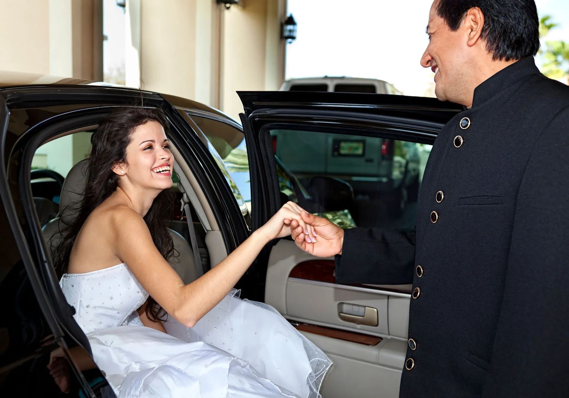 Bride exiting a black car, helped by a man in a black suit. Smiling, outdoors.