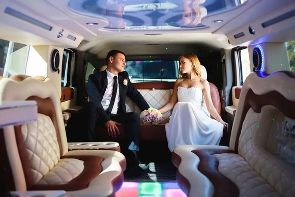 Bride and groom in wedding attire sit in a decorated limo, looking at each other.