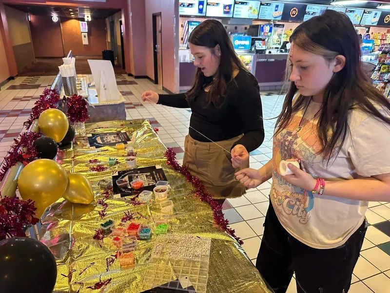 Two girls making jewelry at a decorated table. Gold and black balloons, red tinsel. Theater setting.