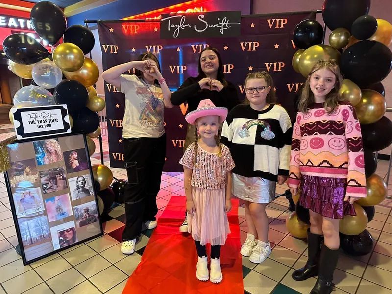 Group of girls at a Taylor Swift-themed VIP event. Red carpet, balloons, and posters. Smiling and posing.