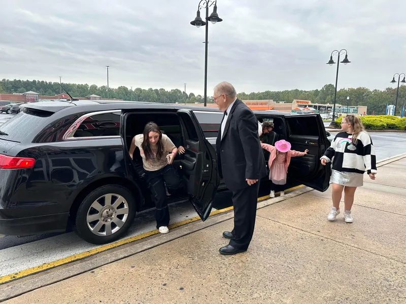 People exiting a black limousine, assisted by a man in a suit, in a parking lot.