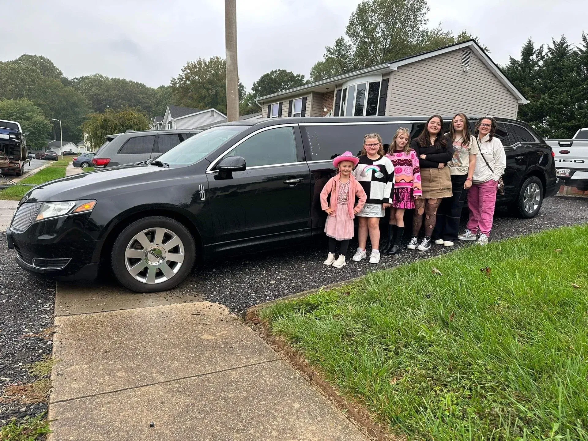Group of girls posing next to a black limousine in front of a house.