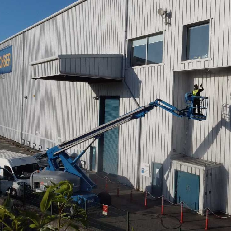 Person in lift bucket working on building exterior. Blue and white building with a utility vehicle parked nearby.