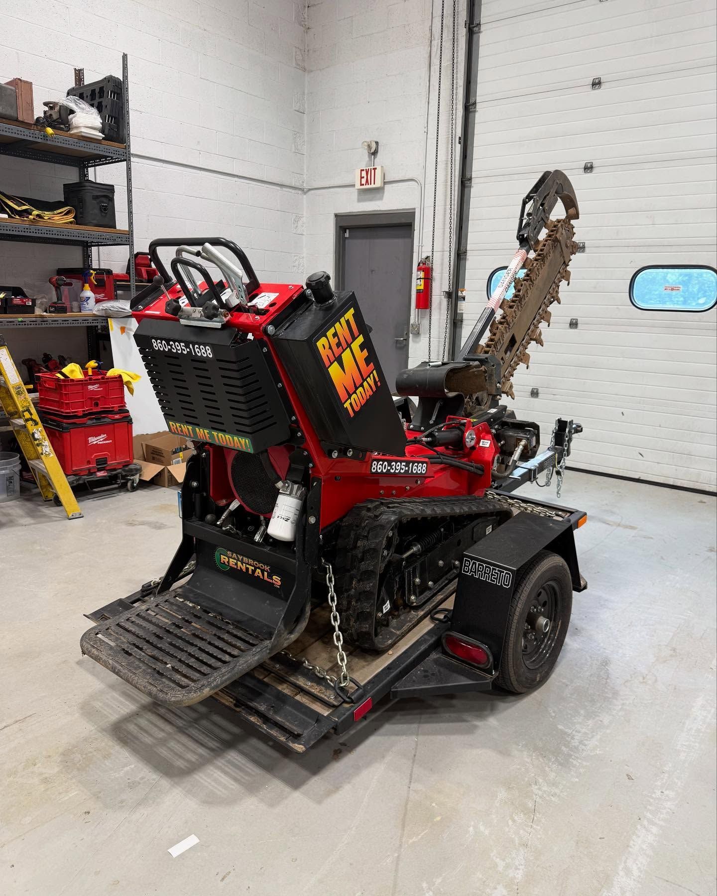 Red trencher on a trailer inside a shop. The machine has a digging chain, black treads, and is ready for use.