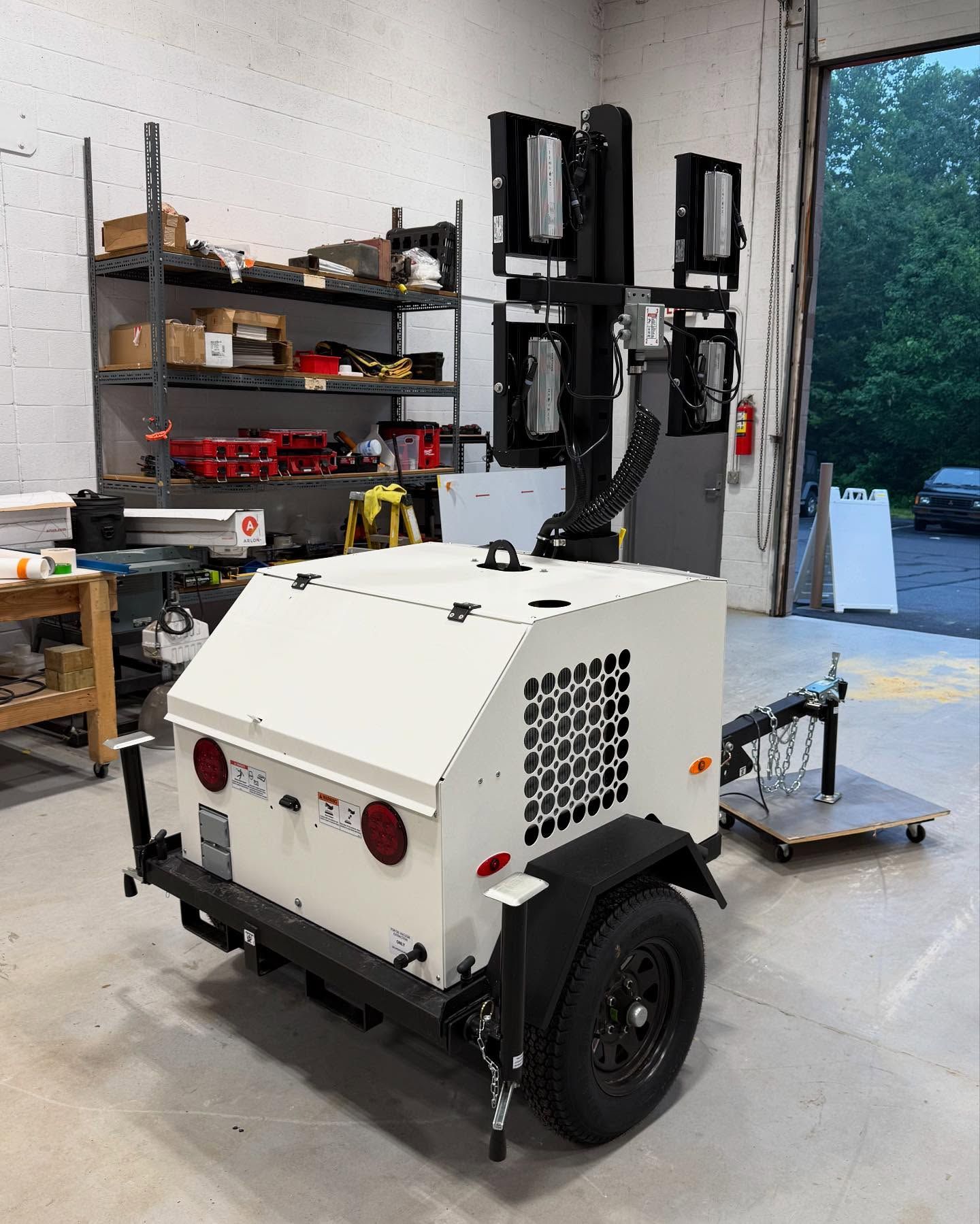 White mobile light tower on a trailer, with four spotlights, parked inside a workshop.