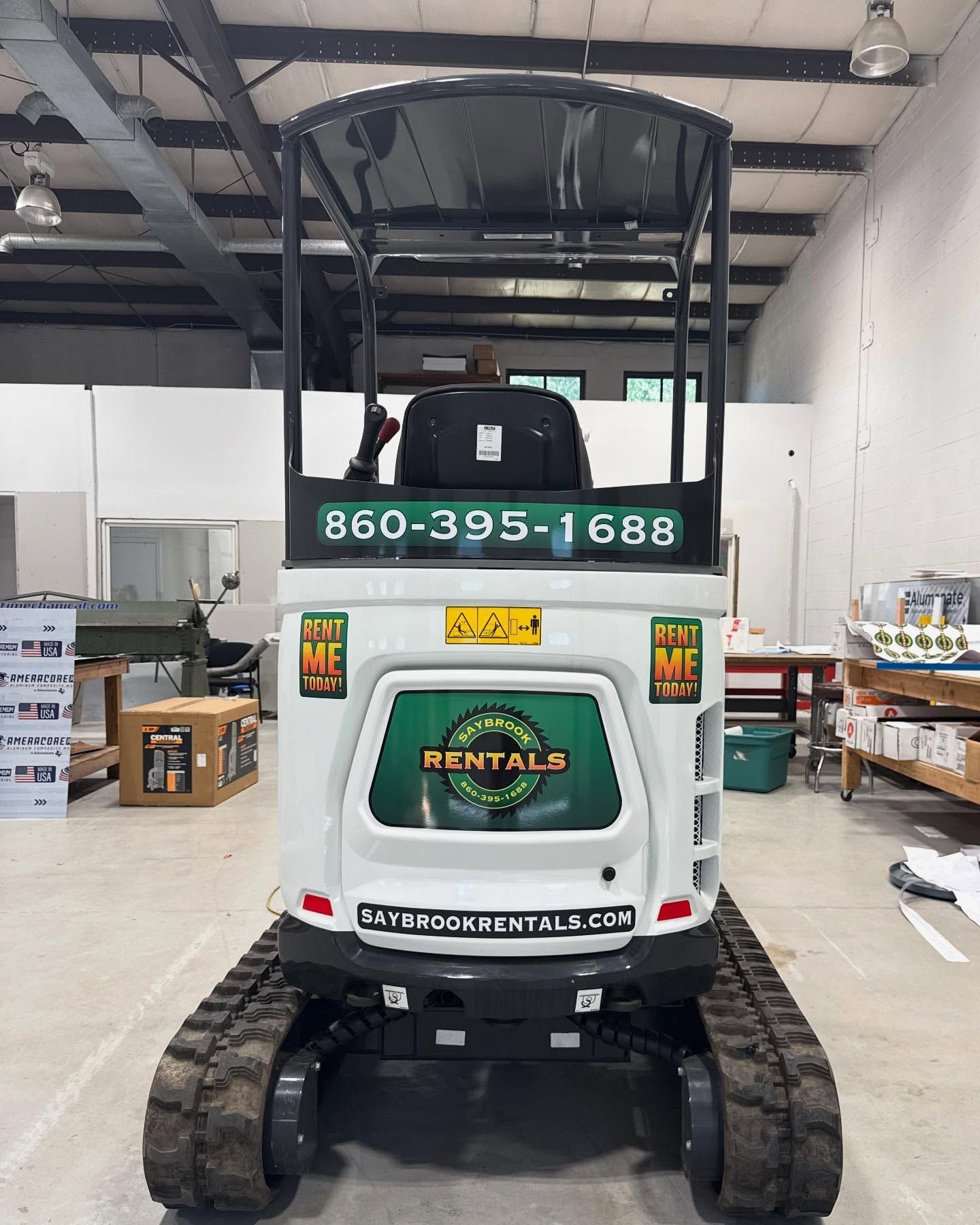 White mini excavator with black tracks and roll cage, with phone number on rear in a warehouse.